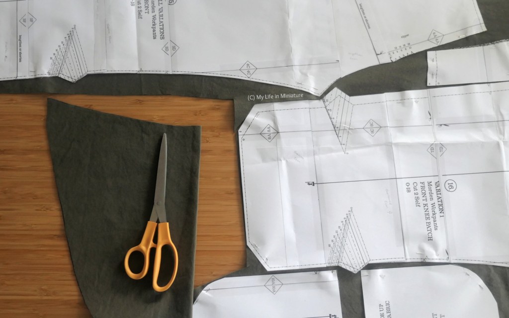 A partially cut-out pant pattern is photographed from above, on a wood surface. Multiple pattern pieces are pinned to the dark green fabric, and a pair of fabric scissors is on the table beside the fabric.
