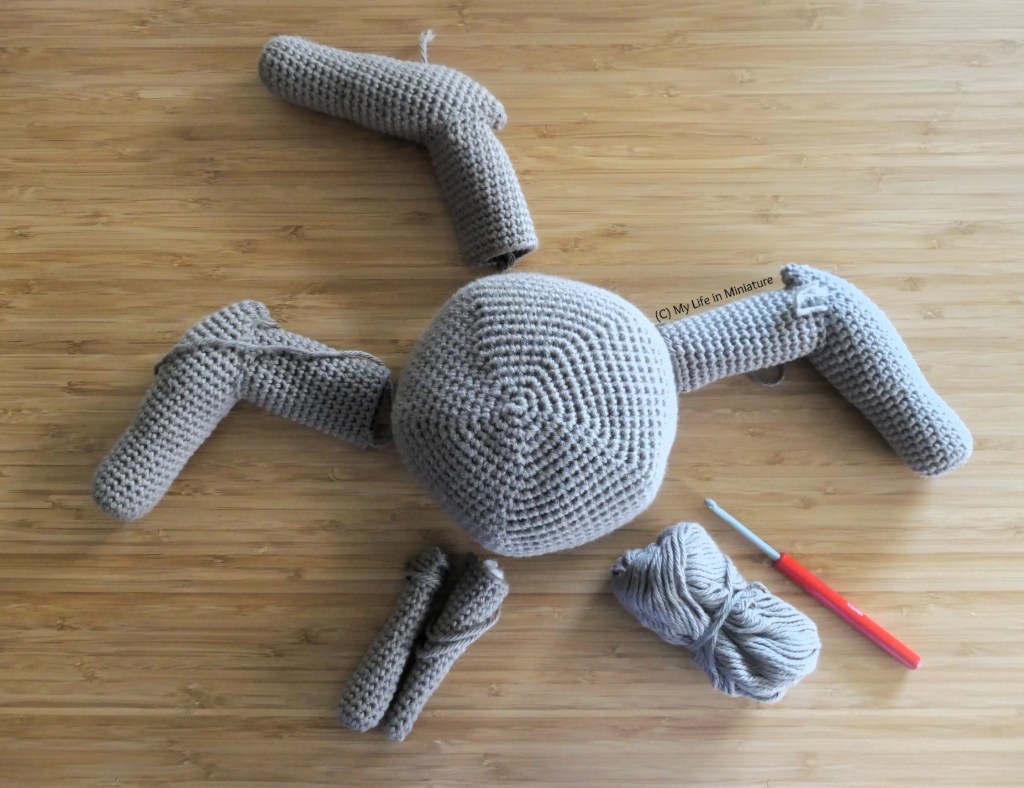 A partially-complete crocheted toy is photographed from above on a wood surface. It looks a lot Rocky from Project Hail Mary, with three complete limbs arranged around a pentagonal body. The other two "limbs" are one incomplete limb, and the yarn and hook the author is using to make the toy.