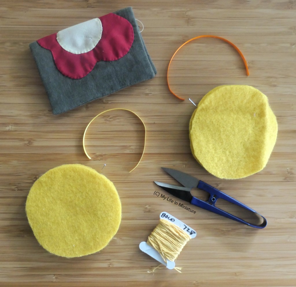 An assortment of sewing supplies, and an unfinished sewing project, are photographed from above on a wood surface. There are two yellow felt circles, with small lengths of ribbon, a needlebook, a card of yellow thread, and a pair of thread snips scattered around pleasingly.