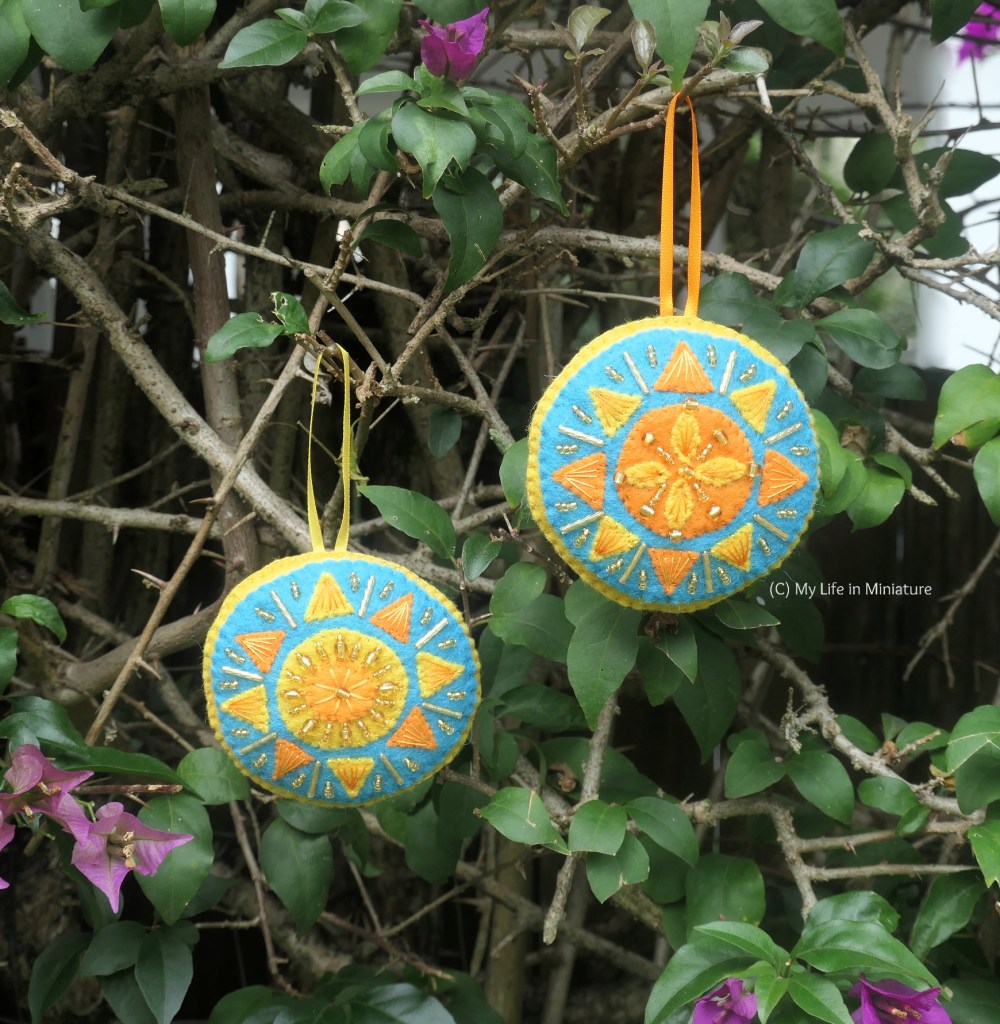 The two sun ornaments hang from the branches of a bougainvillea hedge. One sun ornament has a yellow centre, and the other has an orange centre. Both have alternating yellow and orange triangles surrounding them.