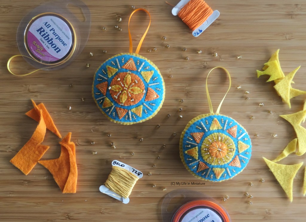 Two completed circular beaded sun ornaments are photographed from above on a wood surface. The suns are on blue backgrounds, and have ribbon hanging loops. Surrounding the ornaments are the supplies used to make them - yellow and orange felt scraps, ribbon, yellow and orange thread, and small gold beads.