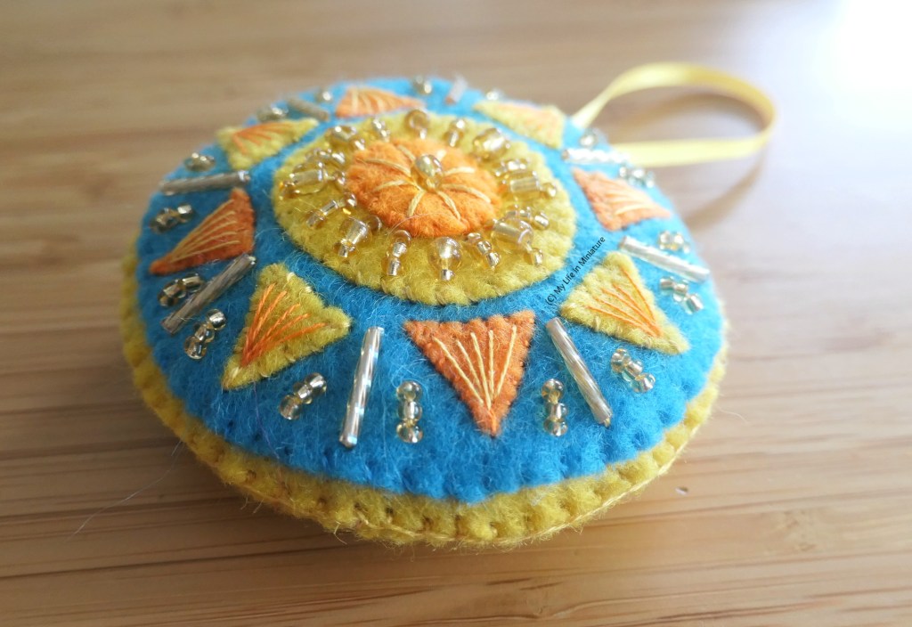 Close-up of the beading and embroidery on one of the sun decorations. The sun is a traditional circle-with-triangles design, with long yellow beads between each triangle. Groups of three small gold beads are between the long beads and the triangle tips.