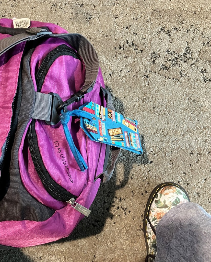 Close-up of the luggage tag tied to the author's purple duffel bag. The bag is photographed from above on a badly-lit airport carpet. The author's shoe is also in the corner of the frame.