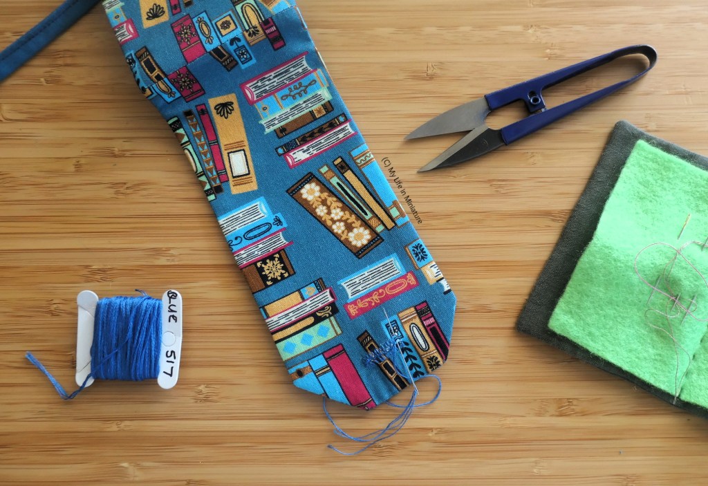 The project is pictured mid-way through having a buttonhole sewn to it. It is surrounded by a spool of embroidery thread, thread snips, and a needle-book.