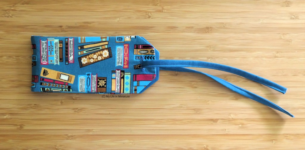 The complete luggage tag is photographed on a wood surface. It's made of blue fabric printed with books, and has a thin blue fabric strap threaded through the top. The tag folds in half to enable the strap to go through (and conceal personal information).