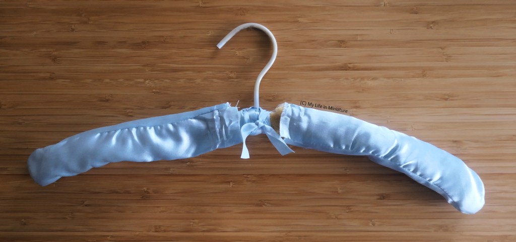 A padded coathanger is photographed from above on a wood surface. It is made of pale blue polyester satin, with a bow around the middle. The raw edges and the degraded foam are sticking out of the middle as well.