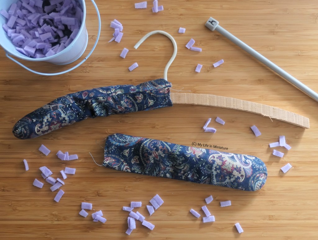 The padded coathanger is photographed from above on a wood surface. It now has one end covered with fresh satin fabric, partially stuffed. Around it are small scraps of fleece, a small bucket filled with said fleece scraps, a knitting needle, and the second end's cover.