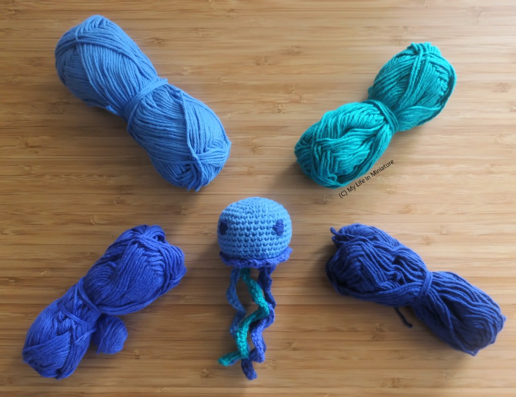 A small crocheted jellyfish stuffed toy is photographed from above on a wood surface. It is mostly blue, and is surrounded by the four balls of yarn used to make it. (They are blue, sea green, navy, and purple.)