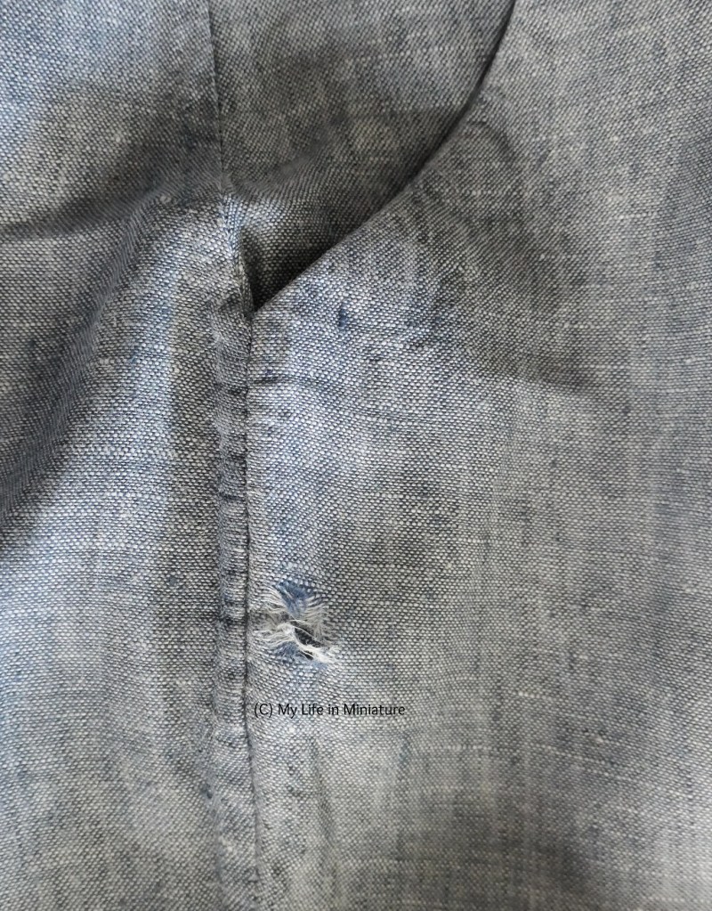 Close-up of a pair of the author's grey-blue handmade pants. The line of the pocket opening is visible at the top of the image, and a small distance below it is a hole, worn through the outer fabric of the pocket.