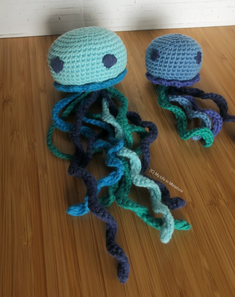 The small crochet jellyfish is photographed next to a larger, paler blue counterpart. Both jellyfish have similar tentacles, embroidered eyes, and frills at the base of their body. The smaller jellyfish is about half the size of the larger one.