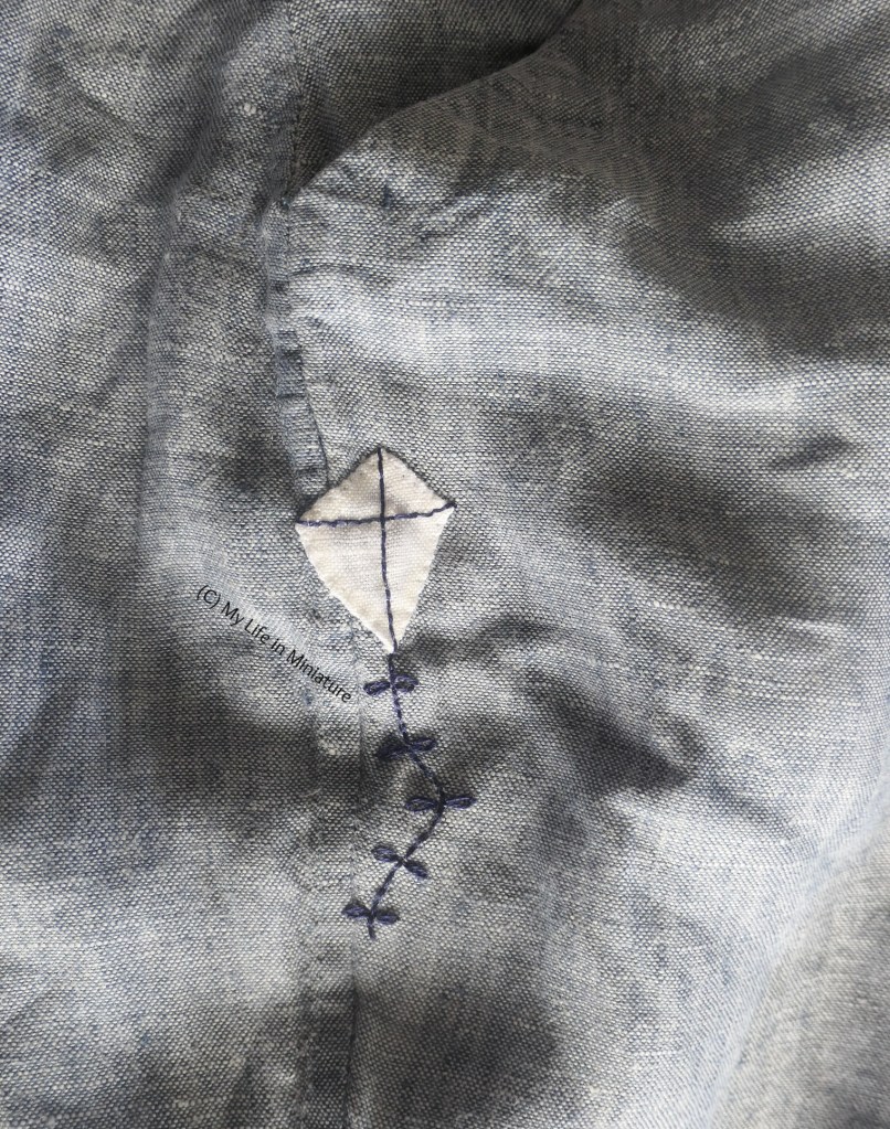 Close-up of the same area of the author's grey-blue pants, below the pocket. The frayed hole has been covered with a patch made to look like a kite. The sail of the kite is white, and the frame and tail are embroidered in navy.