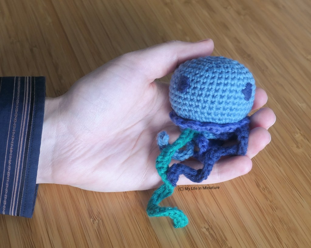 The small jellyfish sits in the author's hand, against a wood background. Its body is small enough to fit in the author's palm.