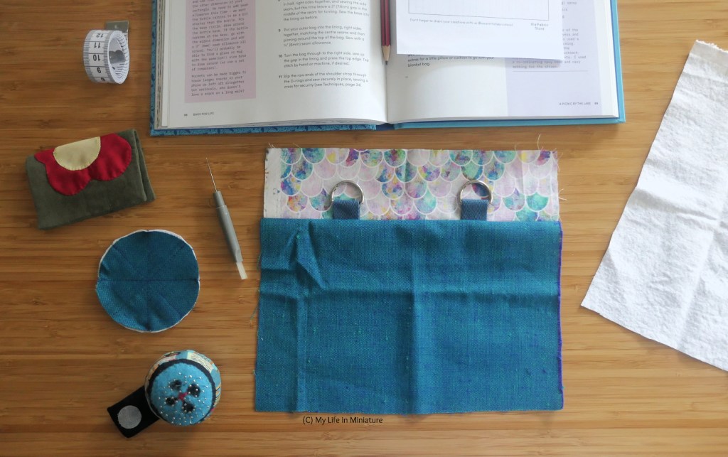 A sewing project is laid out on a wood surface, photographed from above. At the centre is a rectangle of turquoise fabric, with a patterned fabric and two D-rings attached to it. Surrounding this piece of fabric is an open book, a piece of cotton fleece, a pin cushion, an unpicker, a needle-book, and a tape measure.