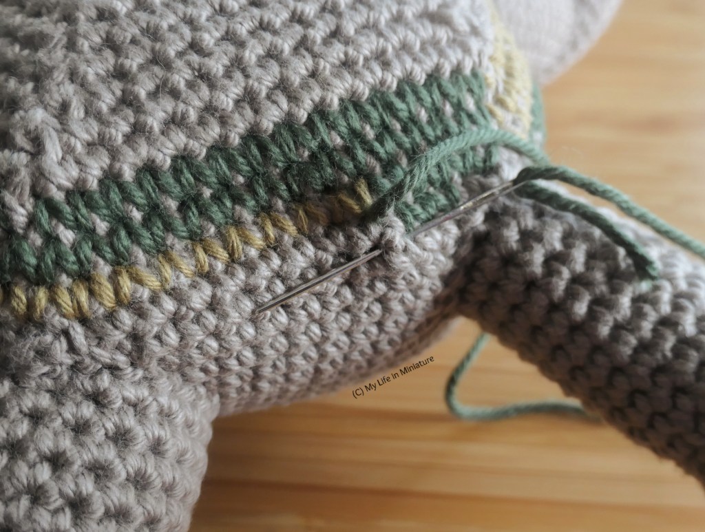 Close-up of a stuffed toy crochet project. The toy is made in brown yarn, and is being embroidered with dark green yarn; a yarn needle is in one of the stitches.