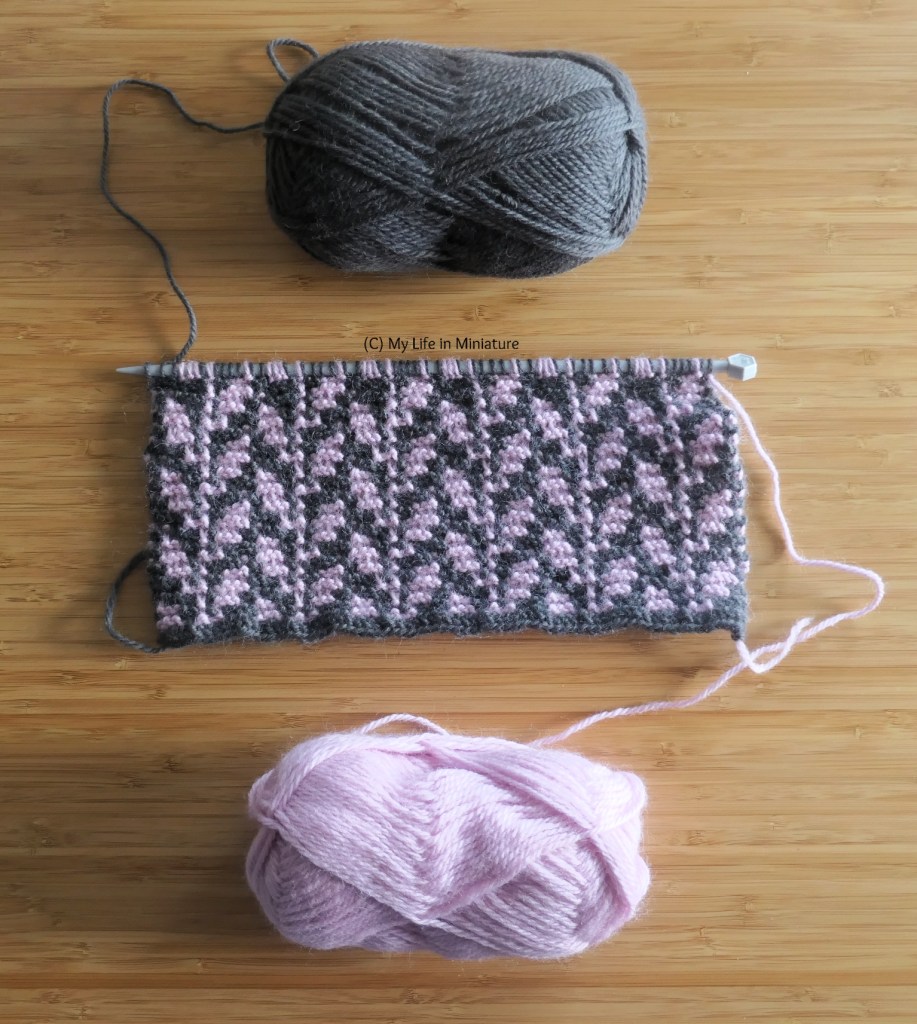 An in-progress knitted scarf is photographed from above on a wood surface. The pattern is of climbing leafy stems, with the leaves in pale pink and the background in dark grey. The corresponding balls of yarn are above and below the knitting.