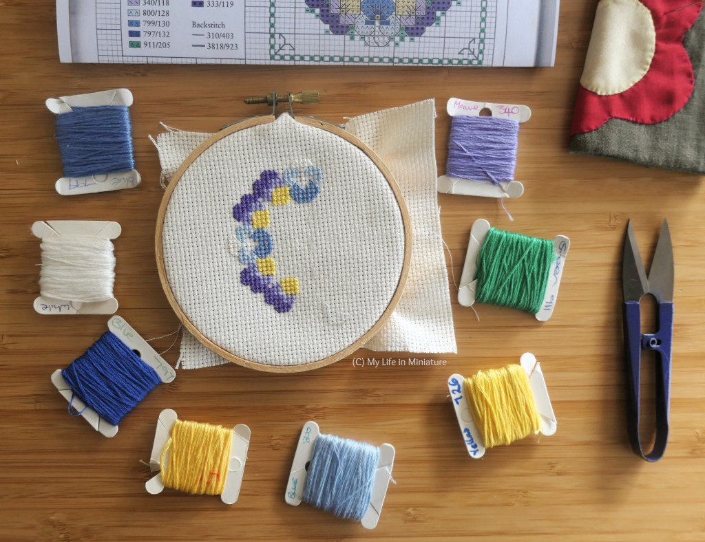 An embroidery hoop is set up on a wood surface with Aida fabric in. It is surrounded by a variety of embroidery threads, thread snips, a needlebook, and a cross-stitching pattern. The hoop has some partially-completed cross-stitching in it, in a rough half-circle shape.