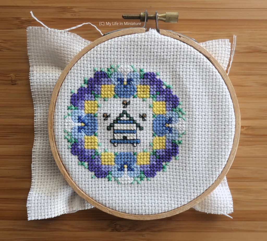 Close-up of an embroidery hoop, fitted with cross-stitching fabric and on a wooden background. A wreath of pansies with a beehive in the middle has been stitched onto the fabric. The pansies are a bit blurred though; only the beehive has had backstitching to define it.