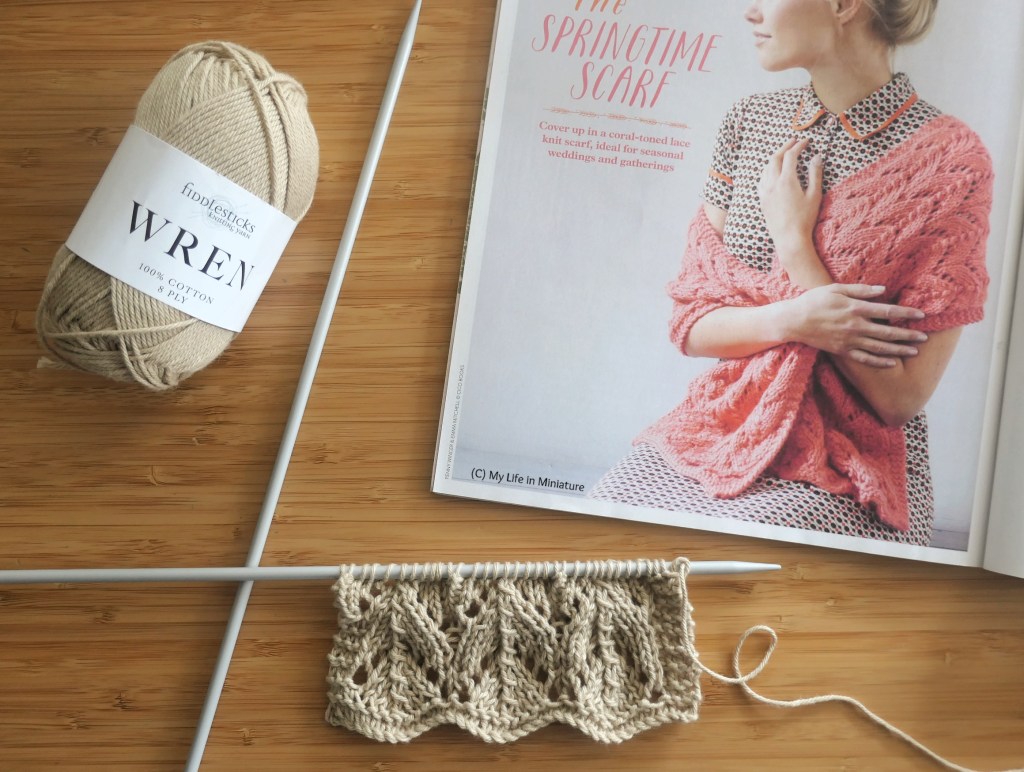 A magazine, open to a knitted scarf pattern, is laid flat on a wood surface. It is surrounded by needles with a sample swatch of the scarf pattern on, and an unopened ball of yarn. The active yarn is very light tan, and the scarf in the magazine picture is coral pink.