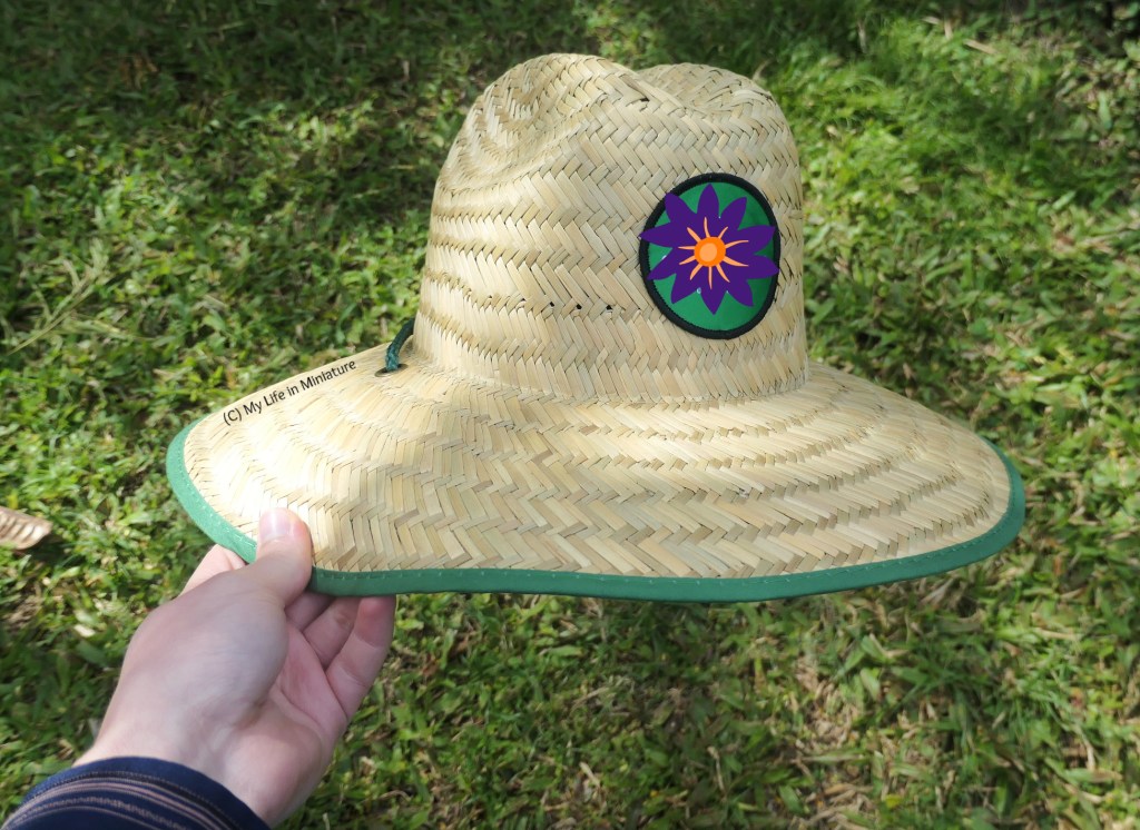 The author's hand holds a straw gardening hat over a grass lawn background. The hat has a dark green cord, barely visible at the back, green trim around the brim, and a circle at centre front with a logo. Said logo on the front of the hat has been covered with a purple flower graphic.