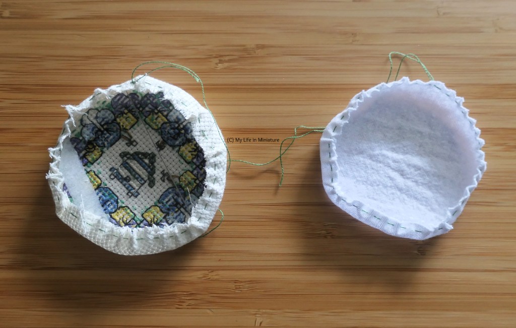Close-up of two fabric circles on a wood background. The left one is the back of the pansy wreath cross-stitching, and the right one is white cotton fleece. Both circles have partially-tightened gathering stitches around the circumference, and clear plastic circles inside them.