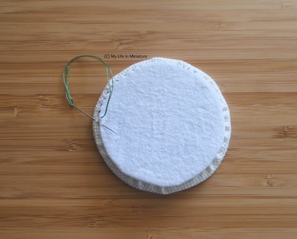 Close-up of the two fabric circles, on a wood surface. The cotton fleece circle is being stitched to the back of the cross-stitching circle with green embroidery thread.