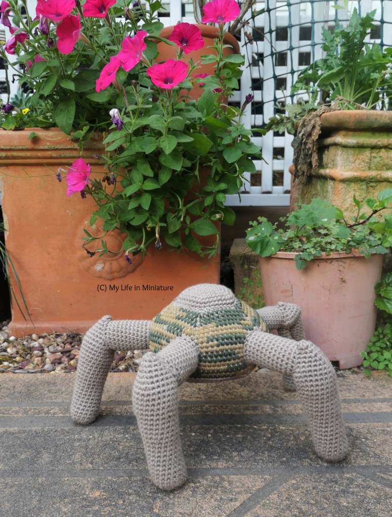 Crochet Rocky is photographed in front of a tall terracotta flower pot, filled with pink petunias.