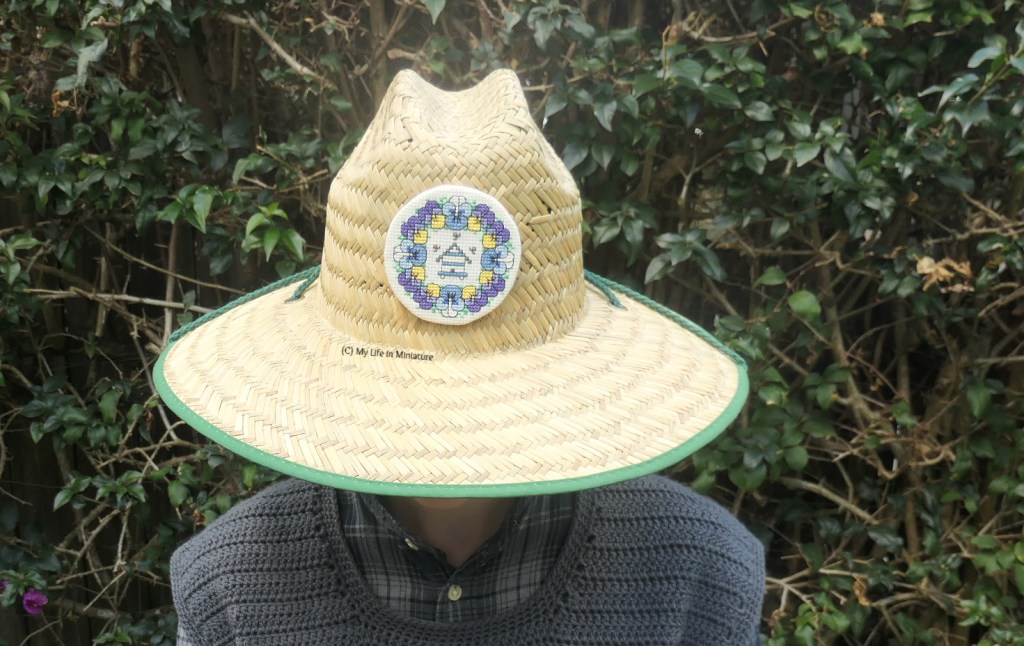 The author is wearing the straw hat from earlier, standing in front of a hedge. The hat's logo on the centre front has been removed and replaced with the complete cross-stitched pansy wreath.  