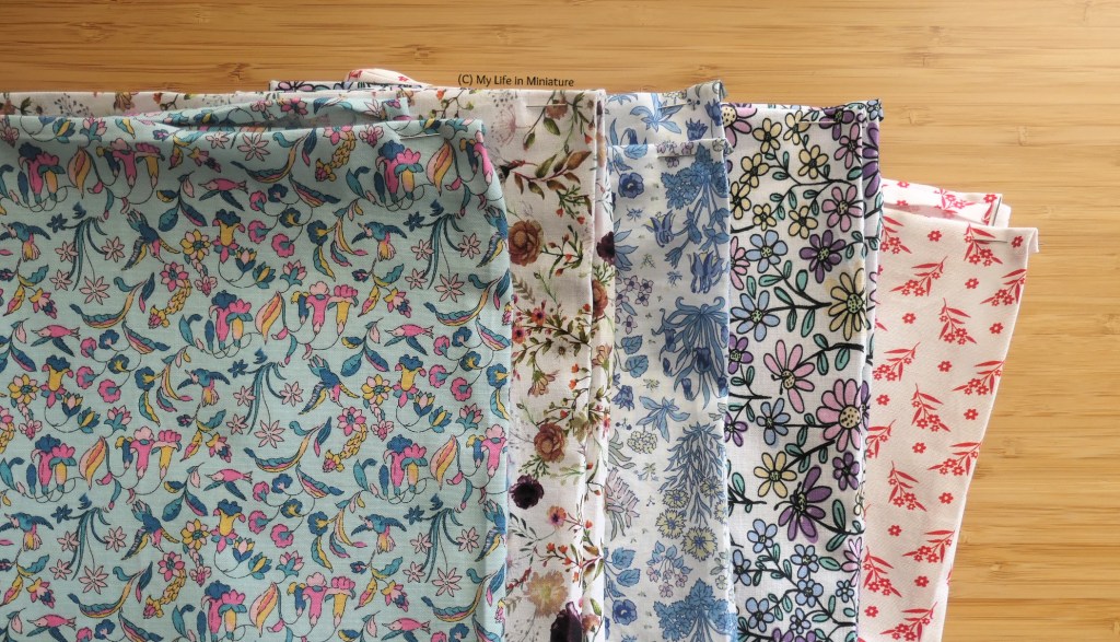 A staggered stack of folded floral cotton fabrics is photographed flat on a wood surface. The top fabric matches the shirt fabric from last image; the four subsequent fabrics are all different. Each fabric has its raw edges ironed in and pinned at the corners.