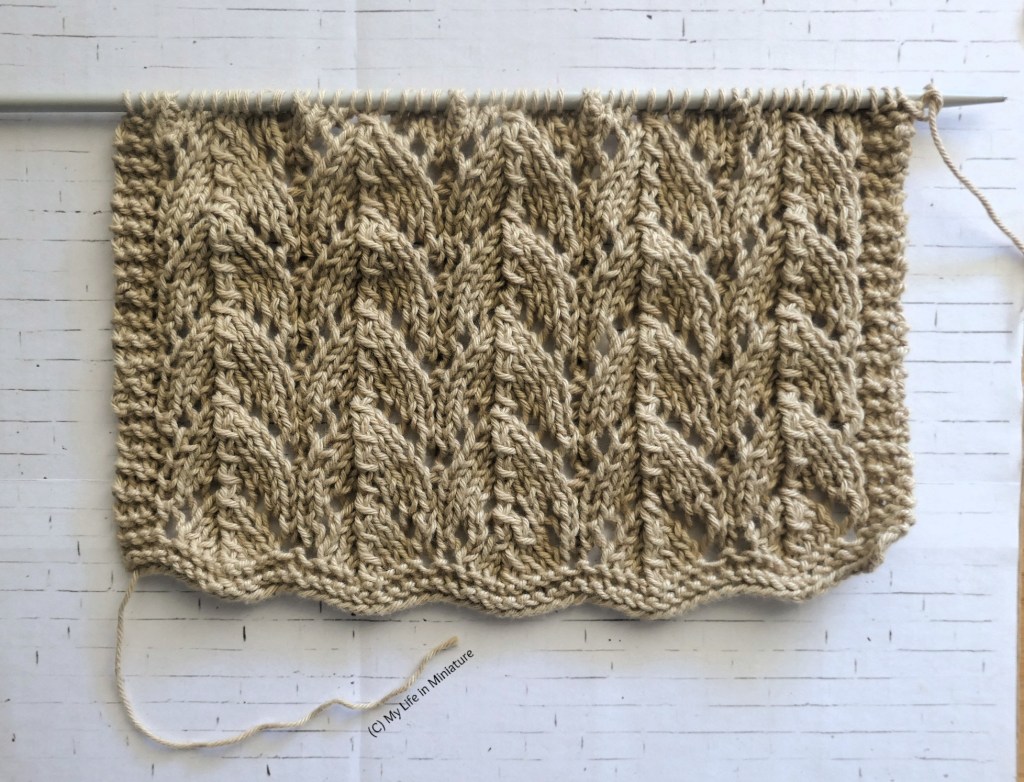 A rectangle of knitting is photographed flat on a white brick background, still on the needle. It is beige, and is a lacy design with a border of plain garter stitch.