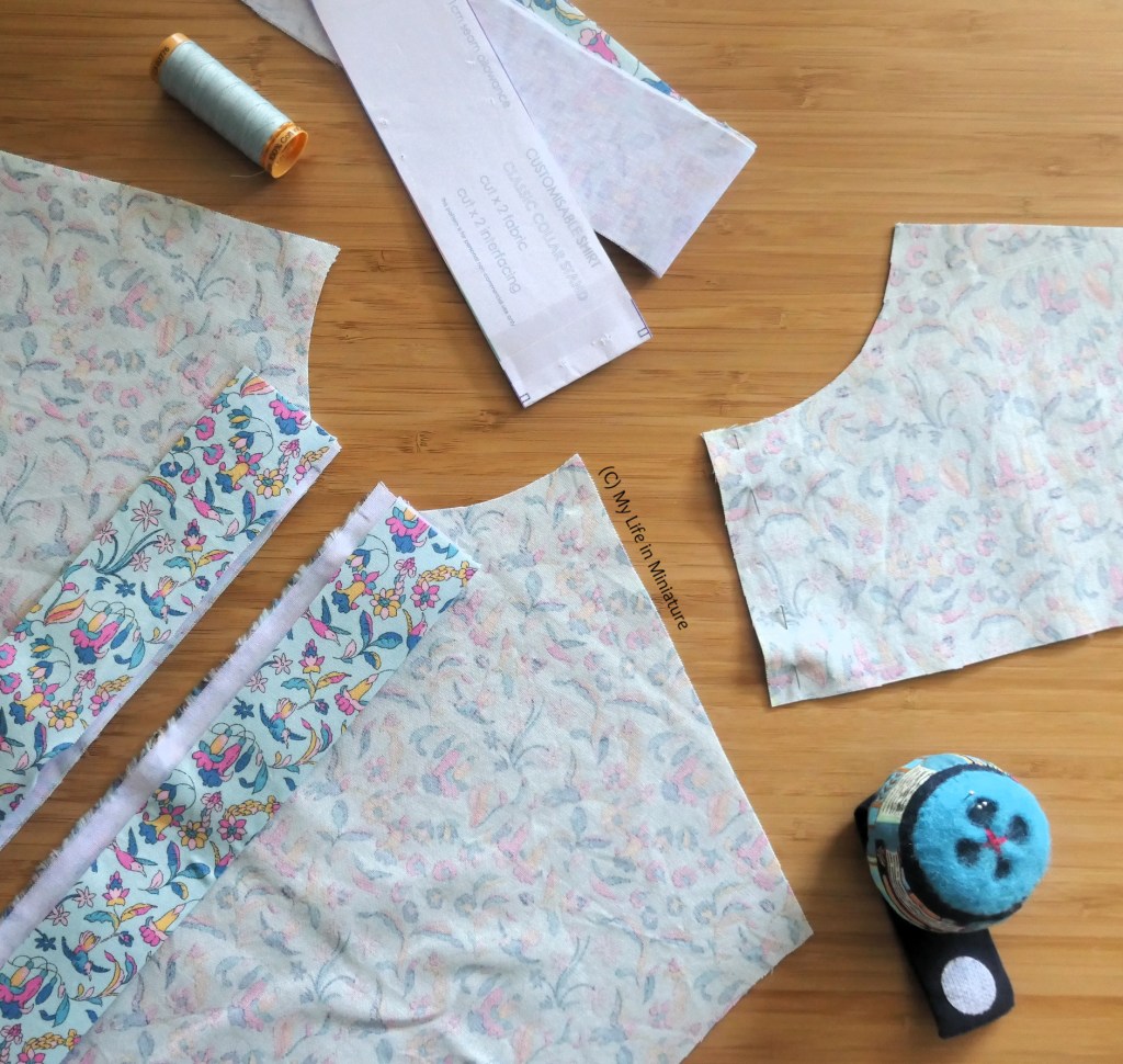 Several fabric pattern pieces for a buttoned shirt are laid flat on a wood background. Some are pinned together in various ways, and they are surrounded by a reel of thread and a pincushion. The fabric is pale blue, with pink and yellow flowers on it.