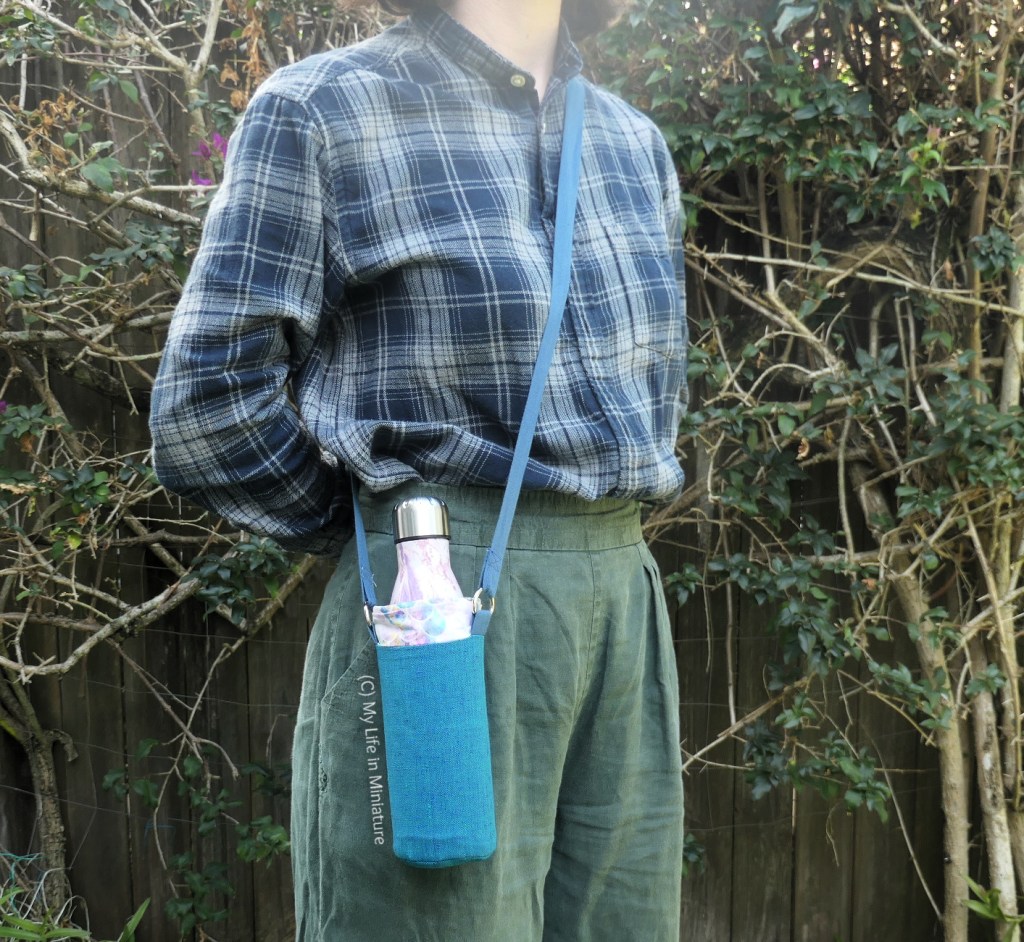 The author stands against a hedge, photographed from neck to thighs. She has a water bottle in a water bottle holder resting against her hip, with the strap over her opposite shoulder. The holder is mostly turquoise, with a small contrast panel of pale purple, and a blue strap. She's wearing a navy checked shirt and green pants.