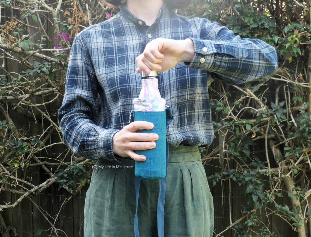The author stands against a hedge, photographed from neck to thighs. She's holding the water bottle in its holder in one hand, and unscrewing the lid with the other. The strap of the holder hangs down beyond the shot. She's wearing a navy checked shirt and green pants.