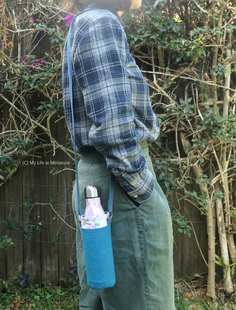 The author stands side-on to the camera against a hedge, photographed from neck to thighs. She has the water bottle holder resting against her side, with the strap over the same shoulder. She's wearing a navy checked shirt and green pants, and has her closest hand in her pocket.
