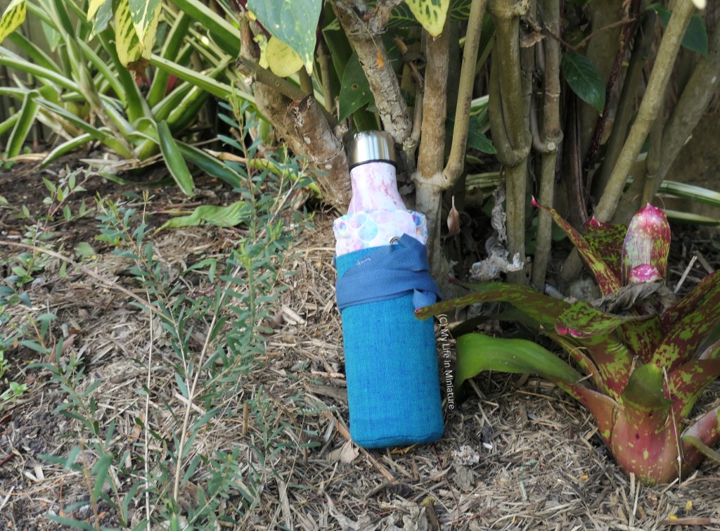 The water bottle in its holder rests on the ground in a garden bed, at the base of a (large) plant. The strap has been wrapped around the bottle and tucked into itself.