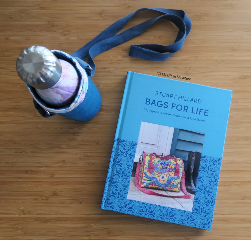 The water bottle in its holder is photographed next to the book it came from, "Bags for Life" by Stuart Hilliard. They are on a wood surface, photographed from above.